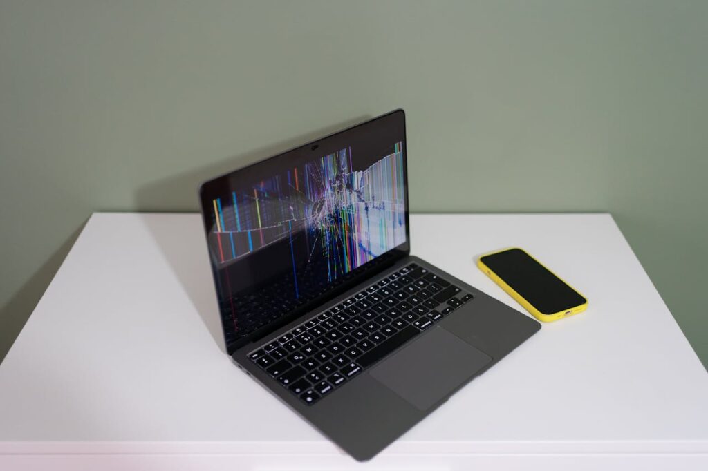 A damaged laptop screen with colorful lines and a yellow smartphone on a white desk.